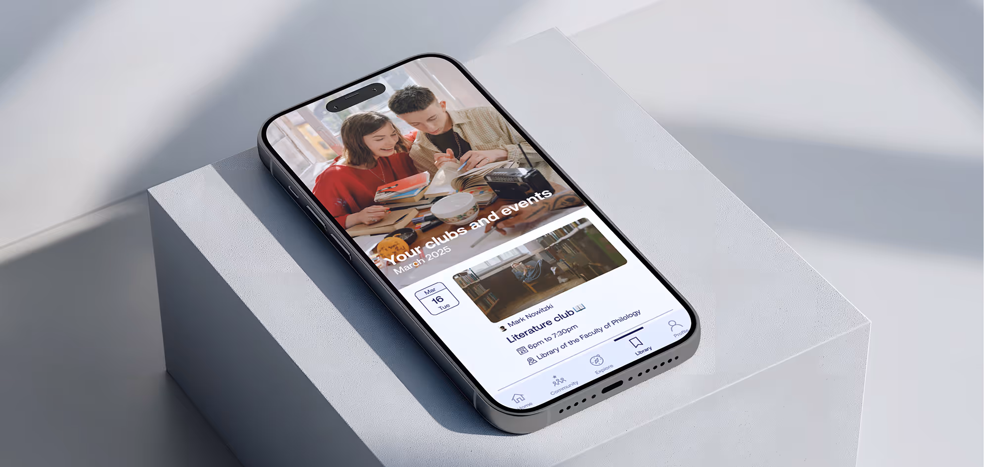 Smartphone on a white pedestal displaying an app page about clubs and events with an image of two people studying and details for a literature club meeting.